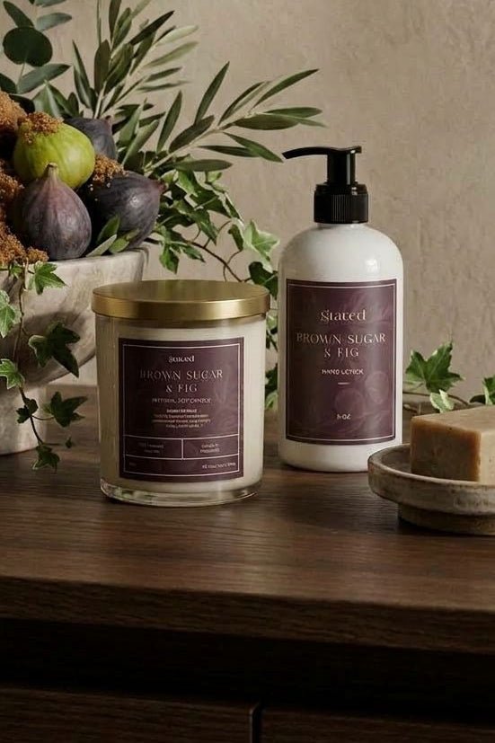A styled bathroom or spa setup on a wooden surface featuring a jar candle with a gold lid, a white pump bottle with a dark label, a soap bar on a small dish, and a reed diffuser with greenery and figs in the background against a textured beige wall.