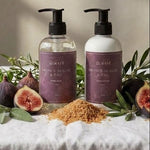 Two pump bottles of Swocet Brown Sugar & Honey body wash and hand wash arranged on a white linen tablecloth with fresh figs, green leaves, and brown sugar piles, against a textured beige wall with soft window light.