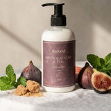 A pump bottle of St. Godeed Brown Sugar & Fig hand wash sits on a white draped tablecloth, surrounded by fresh figs, mint leaves, brown sugar cubes, and rhubarb stems against a textured beige wall with soft window shadows.