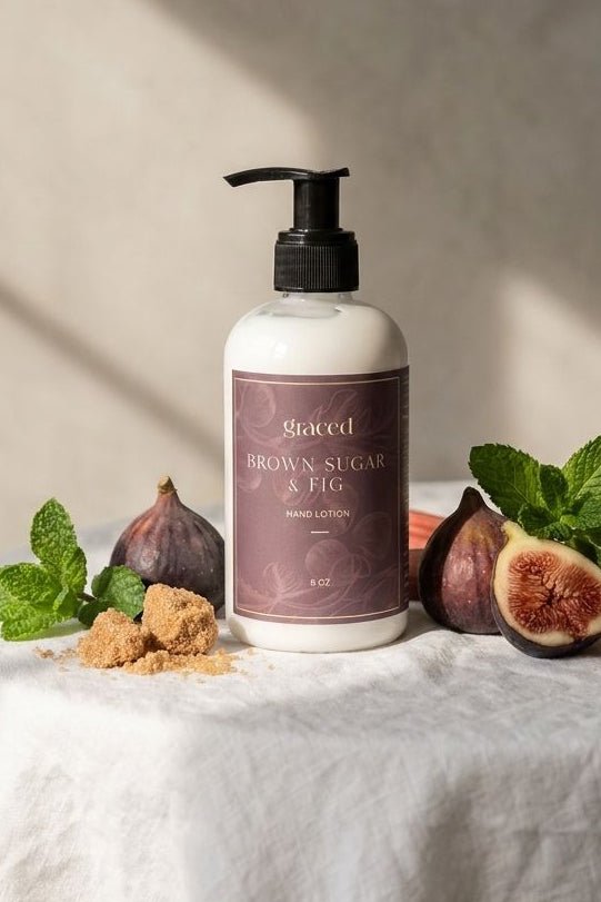 A pump bottle of St. Godeed Brown Sugar & Fig hand wash sits on a white draped tablecloth, surrounded by fresh figs, mint leaves, brown sugar cubes, and rhubarb stems against a textured beige wall with soft window shadows.