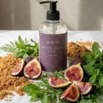 A clear pump bottle labeled 'poached brownsugar & honey' stands on a white cloth, surrounded by fresh figs, mint leaves, green herbs, and brown sugar powder in a softly lit indoor setting.