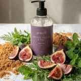 A clear pump bottle labeled 'poached brownsugar & honey' stands on a white cloth, surrounded by fresh figs, mint leaves, green herbs, and brown sugar powder in a softly lit indoor setting.