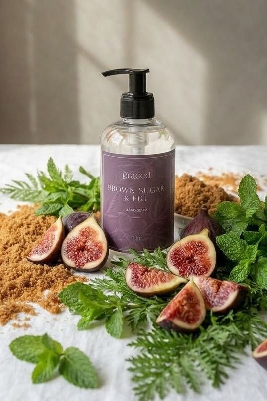 A clear pump bottle labeled 'poached brownsugar & honey' stands on a white cloth, surrounded by fresh figs, mint leaves, green herbs, and brown sugar powder in a softly lit indoor setting.