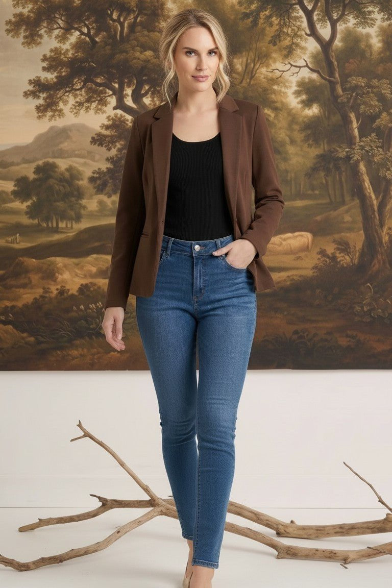 A woman wearing a graced brown knit blazer over a black top and blue jeans, standing in front of a landscape painting.