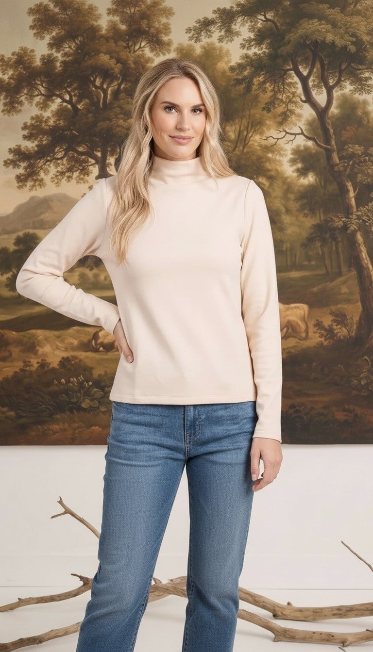 Model wearing a graced ivory long sleeve mock neck knit top paired with blue jeans against a landscape painting backdrop