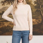 Model wearing a graced ivory long sleeve mock neck knit top paired with blue jeans against a landscape painting backdrop