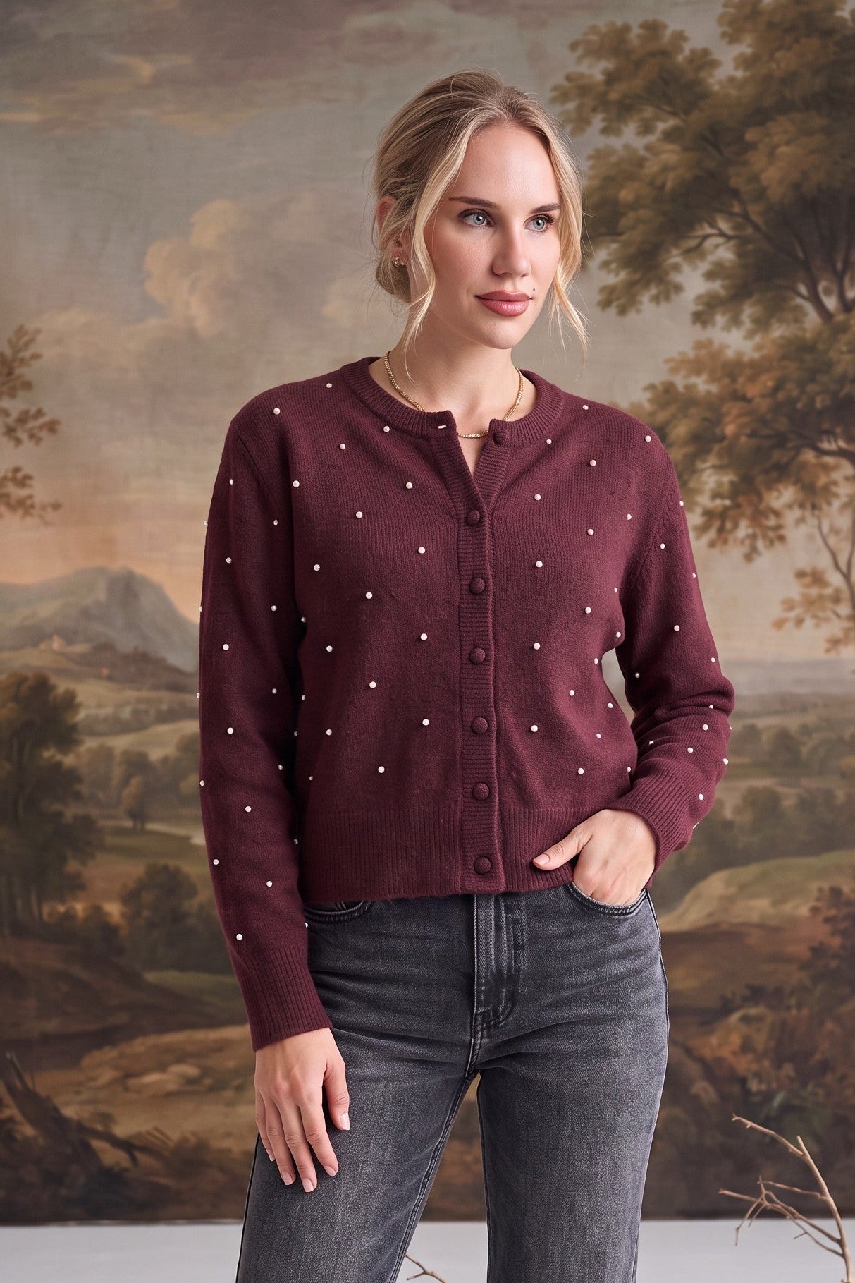 A woman modeling a burgundy button-up cardigan with small white embroidered dots, paired with dark gray jeans, standing in front of a classical landscape painting backdrop.