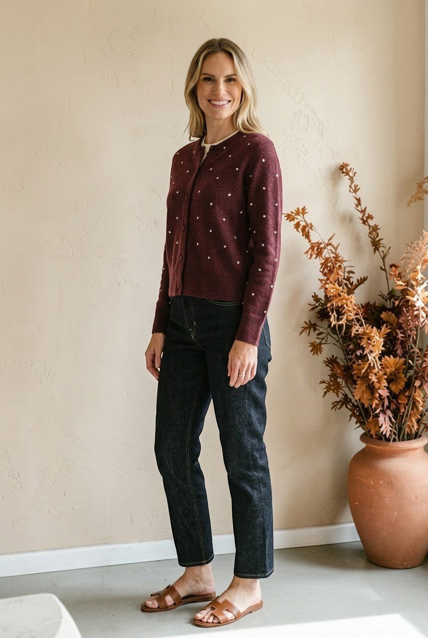 Woman wearing a graced burgundy cardigan with raised white polka dots, styled with dark denim jeans and brown sandals.