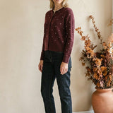 Woman wearing a graced burgundy cardigan with raised white polka dots, styled with dark denim jeans and brown sandals.
