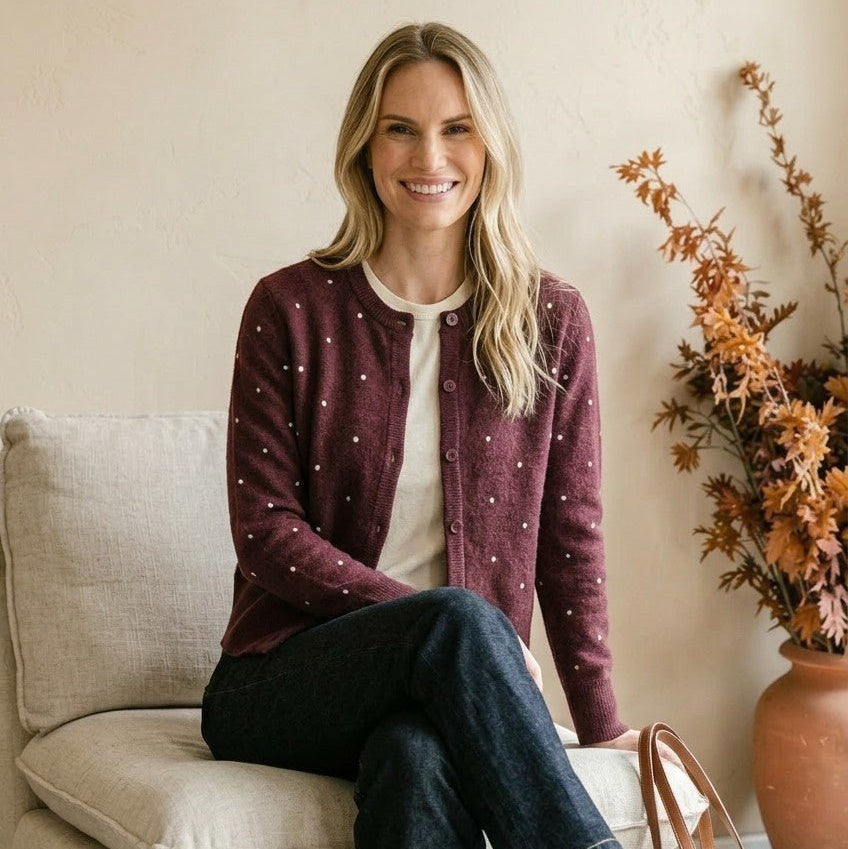 Woman wearing a burgundy graced raised polka dot cardigan over a cream top and dark jeans, sitting on a beige chair.