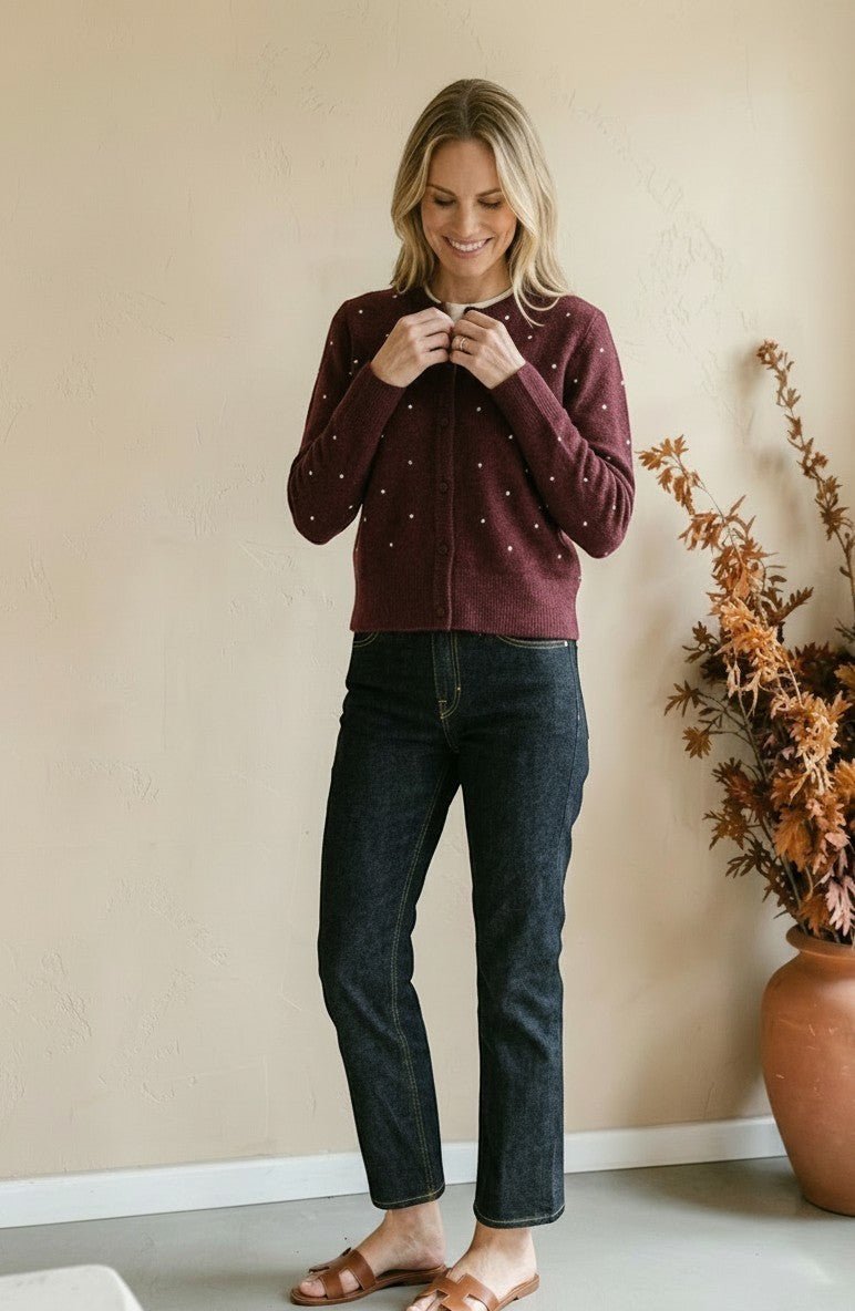 Woman wearing a graced burgundy cardigan with raised white polka dots, paired with dark denim jeans.