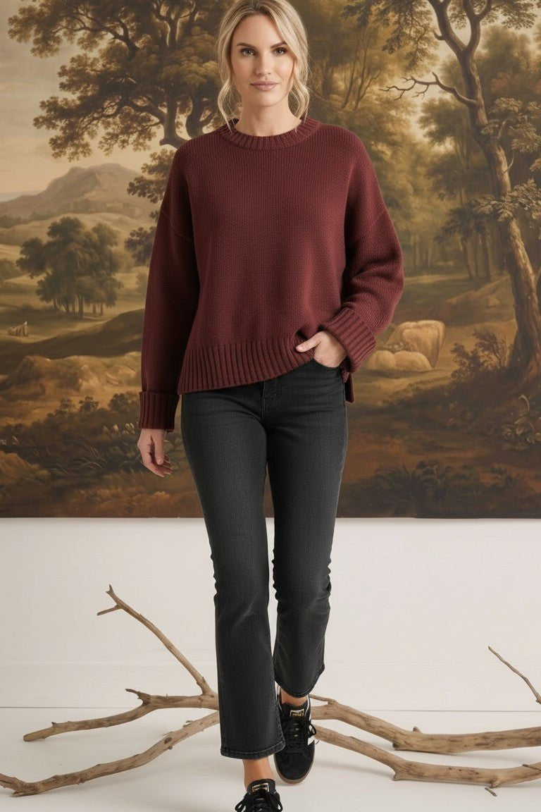Model wearing a graced ribbed cuff sweater in deep coco brown with black jeans against a landscape mural background