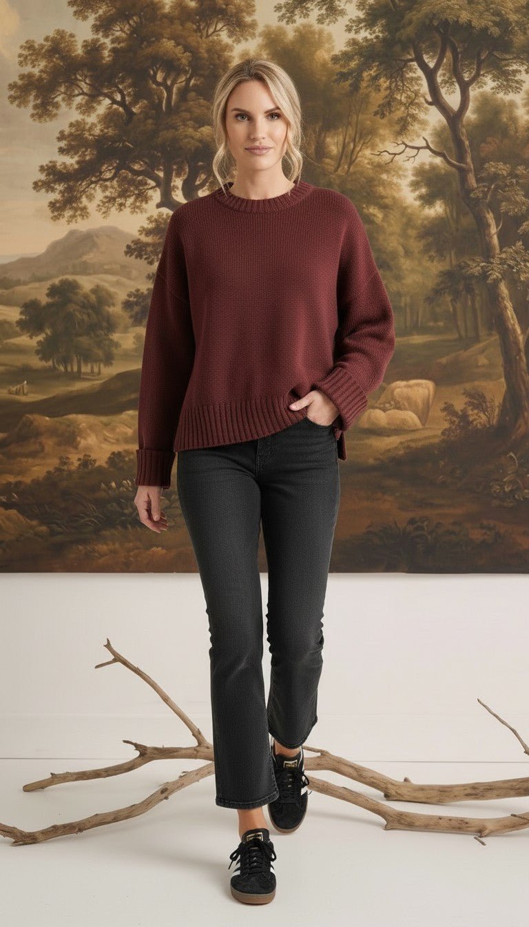 Model wearing a graced ribbed cuff sweater in deep coco brown with black jeans against a landscape mural background
