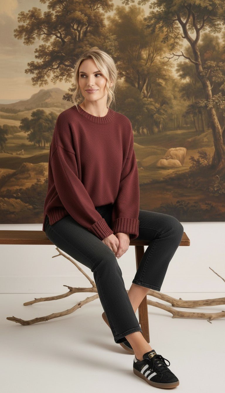 A model wearing a coco-coloured graced ribbed cuff sweater with black jeans, sitting on a wooden bench.