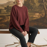 A model wearing a coco-coloured graced ribbed cuff sweater with black jeans, sitting on a wooden bench.