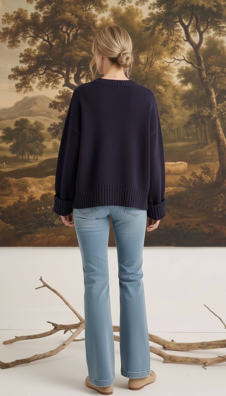 Model wearing a navy graced ribbed cuff sleeve sweater with light wash jeans, back view against a landscape painting.