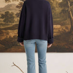 Model wearing a navy graced ribbed cuff sleeve sweater with light wash jeans, back view against a landscape painting.