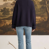 Model wearing a navy graced ribbed cuff sleeve sweater with light wash jeans, back view against a landscape painting.
