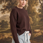 Woman wearing a graced brown crew neck sweater with ribbed cuffs and neckline, styled with light wash jeans.
