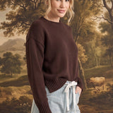 Woman wearing a graced brown crew neck sweater with ribbed cuffs and neckline, styled with light wash jeans.