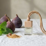 A clear glass perfume bottle with a light wood cap and a curved light wood neck, styled on a white linen tablecloth with figs, mint leaves, and spices in a softly lit interior.