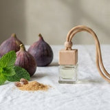 A clear glass perfume bottle with a light wood cap and a curved light wood neck, styled on a white linen tablecloth with figs, mint leaves, and spices in a softly lit interior.