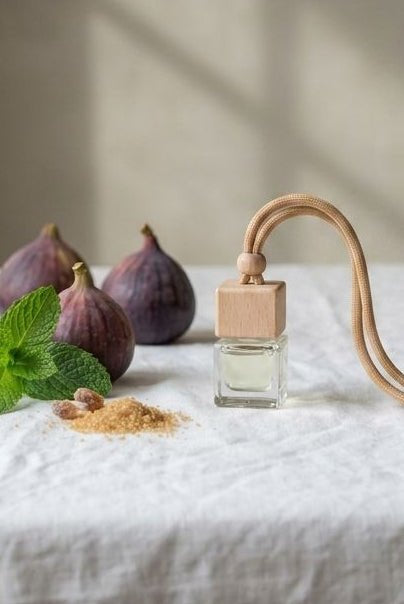 A clear glass perfume bottle with a light wood cap and a curved light wood neck, styled on a white linen tablecloth with figs, mint leaves, and spices in a softly lit interior.