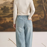 Back view of a woman wearing graced light wash soft denim barrel pants with an elastic waist and patch pockets.