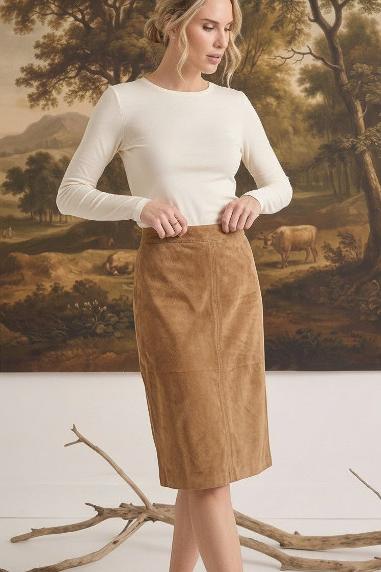 Model wearing a graced tan suede A-line skirt with a long-sleeve cream top in a rustic indoor setting.