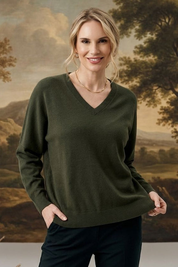 A woman modeling a dark olive green V-neck sweater and black pants, standing in front of a large classical landscape painting.