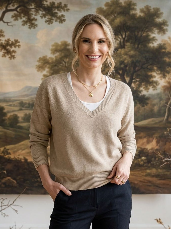 A woman stands indoors wearing a beige v-neck knit sweater over a white top and dark navy pants, with a large landscape painting hanging on the wall behind her.