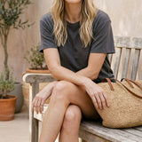 graced model wearing Z Supply The After Hour sunglasses with a dark grey t-shirt and straw tote bag on a wooden bench