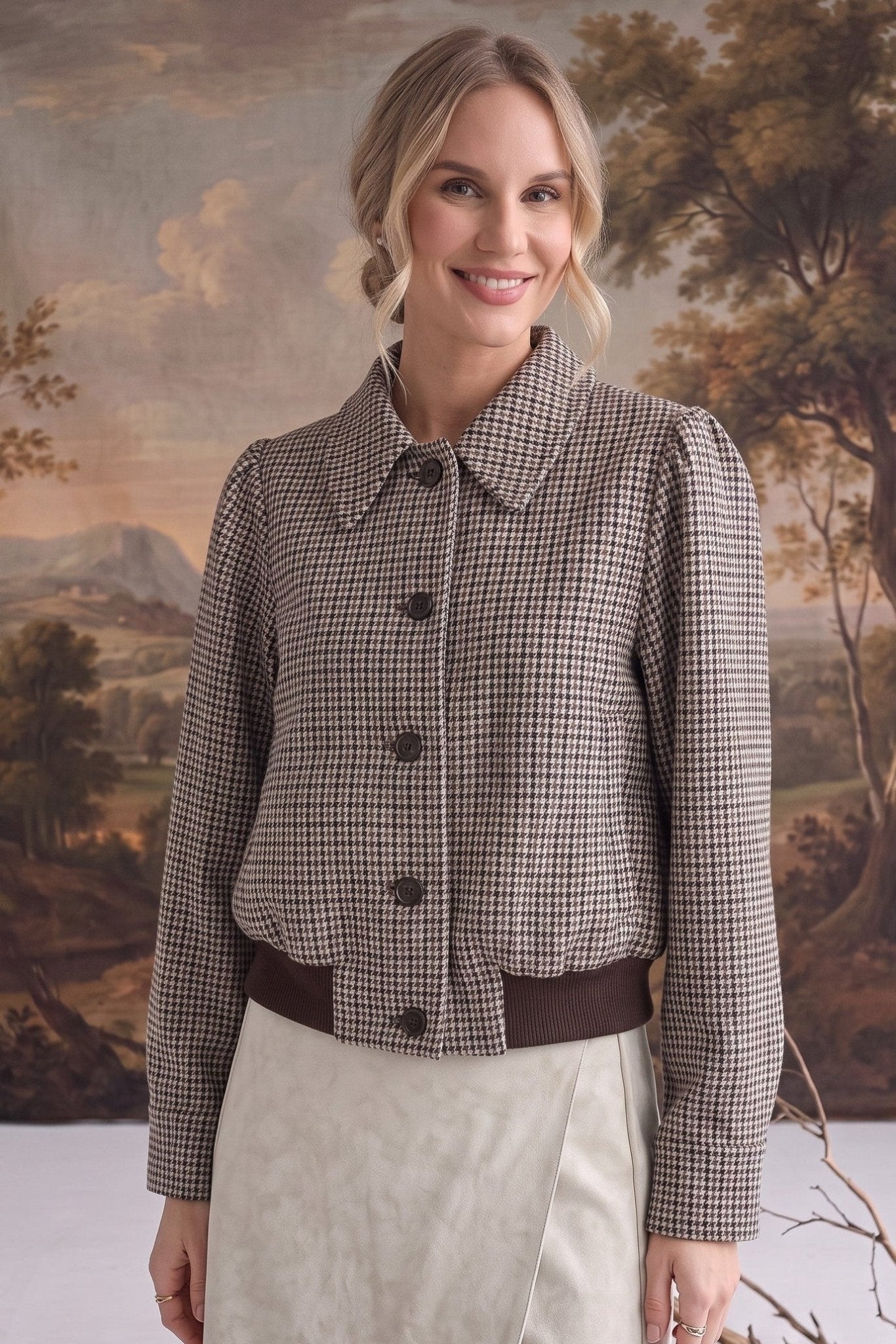 A woman modeling a cropped, long-sleeve button-front jacket with a brown and beige houndstooth pattern, paired with a cream skirt, set against a rustic landscape backdrop.