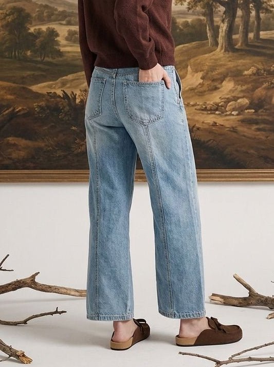 A woman stands in front of a large framed landscape painting, wearing a dark brown long-sleeve top, light blue wide-leg jeans, and brown slip-on shoes.