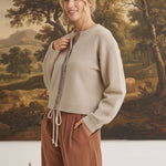 graced Baylie cardigan in beige with gingham trim worn by a blonde model against a landscape painting background