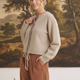 graced Baylie cardigan in beige with gingham trim worn by a blonde model against a landscape painting background