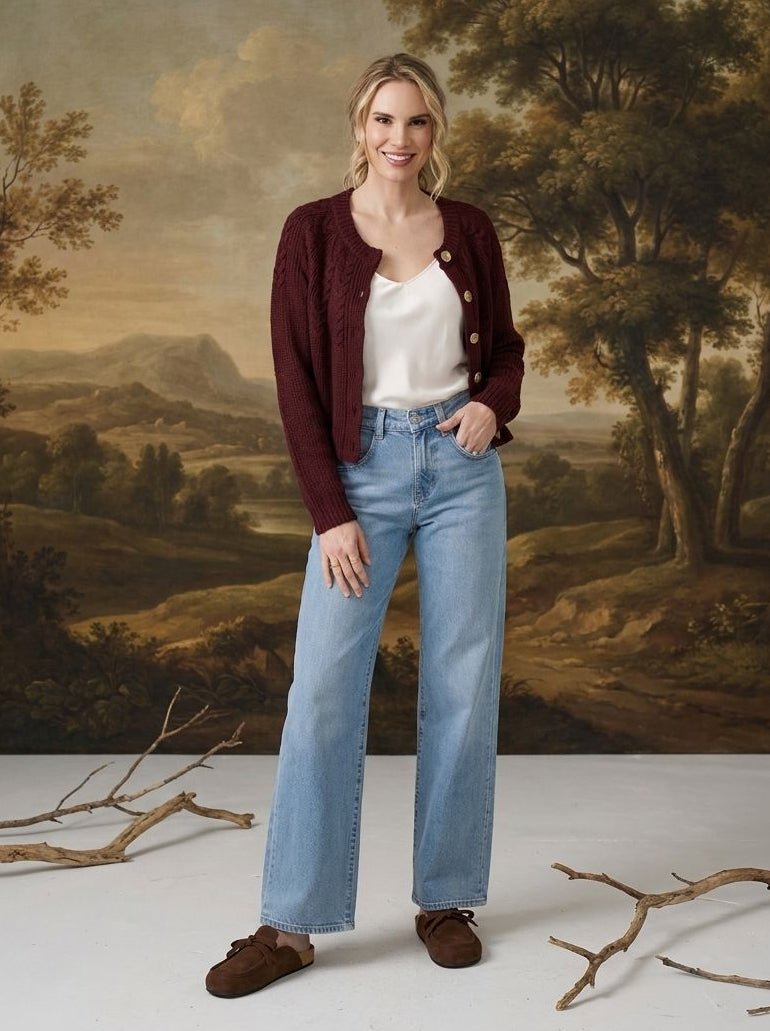 A woman stands in front of a classical landscape painting backdrop, wearing a dark burgundy cardigan over a white top, light blue wide-leg jeans, and brown suede shoes.