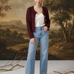 A woman stands in front of a classical landscape painting backdrop, wearing a dark burgundy cardigan over a white top, light blue wide-leg jeans, and brown suede shoes.