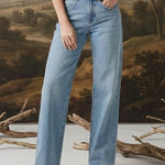 A woman stands in a studio wearing a dark floral puff-sleeve blouse and light blue wide-leg jeans, posed in front of a large classical landscape painting backdrop with decorative branches on the floor.