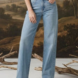 A woman stands in a studio wearing a dark floral puff-sleeve blouse and light blue wide-leg jeans, posed in front of a large classical landscape painting backdrop with decorative branches on the floor.