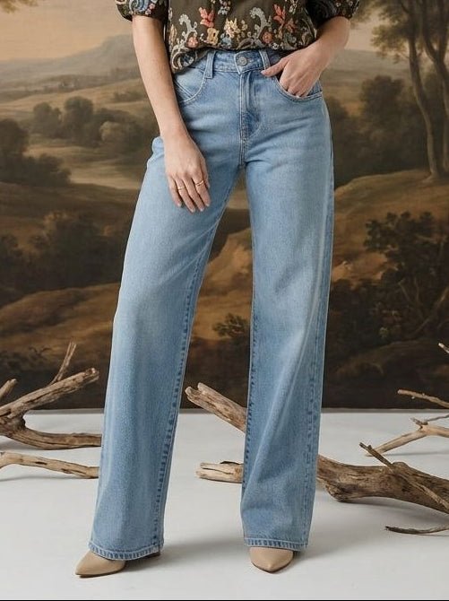 A woman stands in a studio wearing a dark floral puff-sleeve blouse and light blue wide-leg jeans, posed in front of a large classical landscape painting backdrop with decorative branches on the floor.