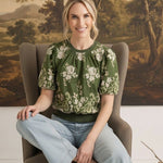 graced Brenda floral top in olive green with puff sleeves and embroidered details worn by a woman in a chair