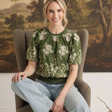graced Brenda floral top in olive green with puff sleeves and embroidered details worn by a woman in a chair