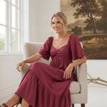 A woman wearing a burgundy graced Celeste puff sleeve midi dress while sitting in a chair in a bright room.