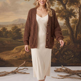 Model wearing a graced mocha chunky knit cardigan with buttons and pockets over a cream slip dress in a studio setting