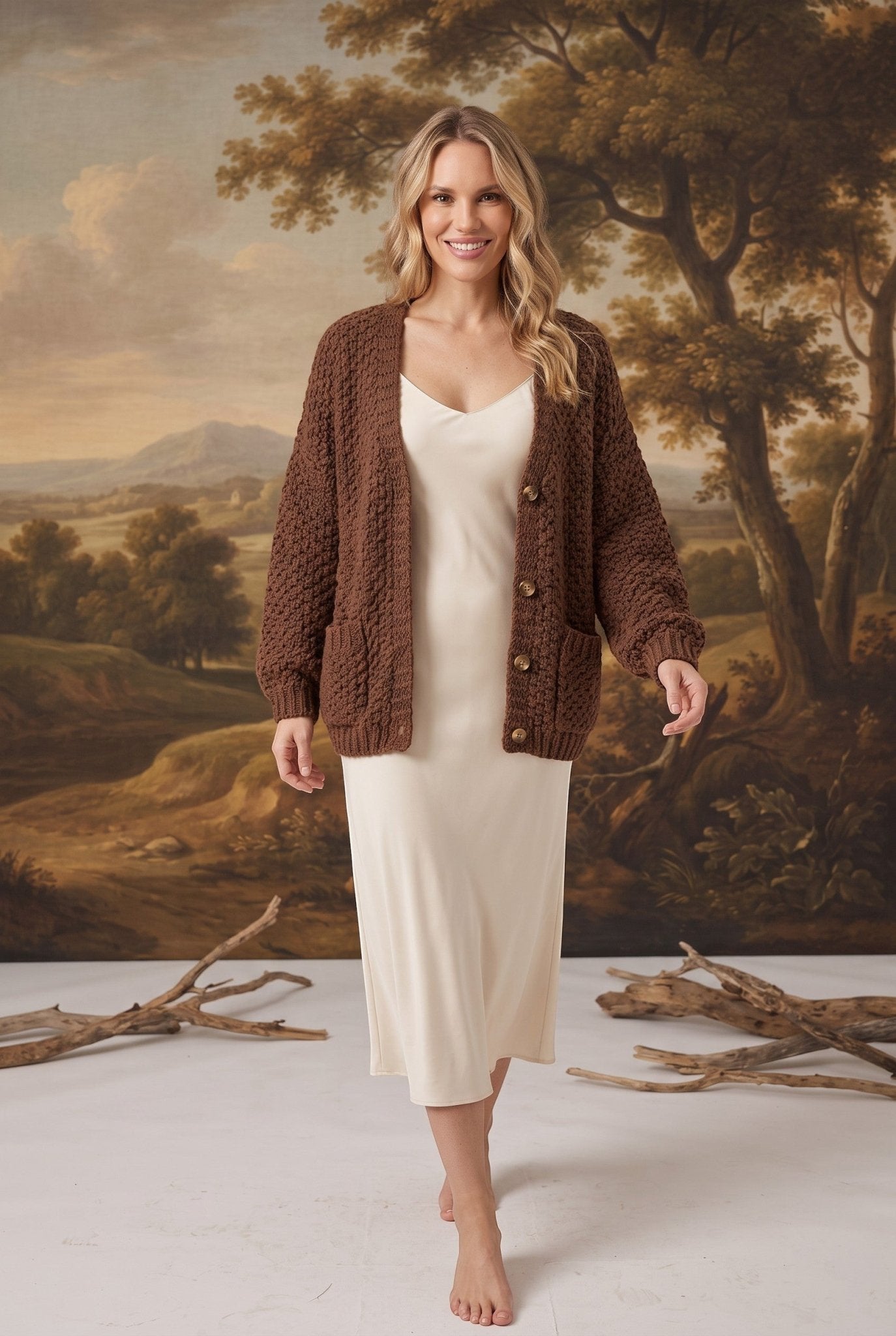 Model wearing a graced mocha chunky knit cardigan with buttons and pockets over a cream slip dress in a studio setting
