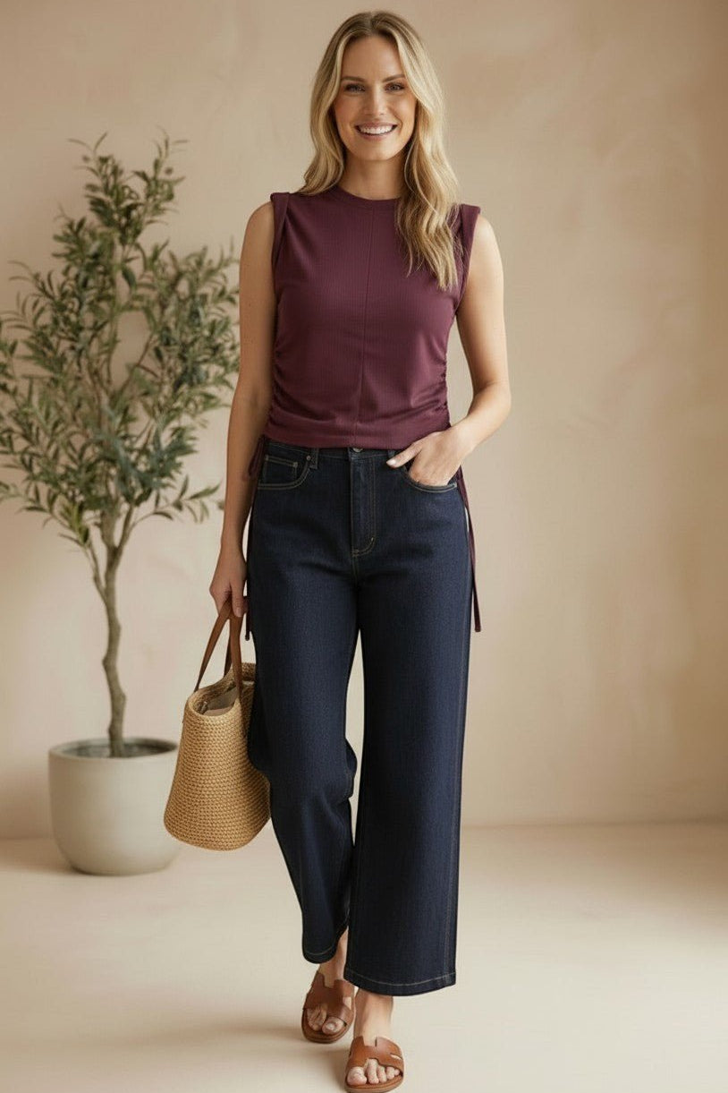 A woman wearing a graced winetasting cotton top with side tie detail and dark denim jeans in a studio setting.