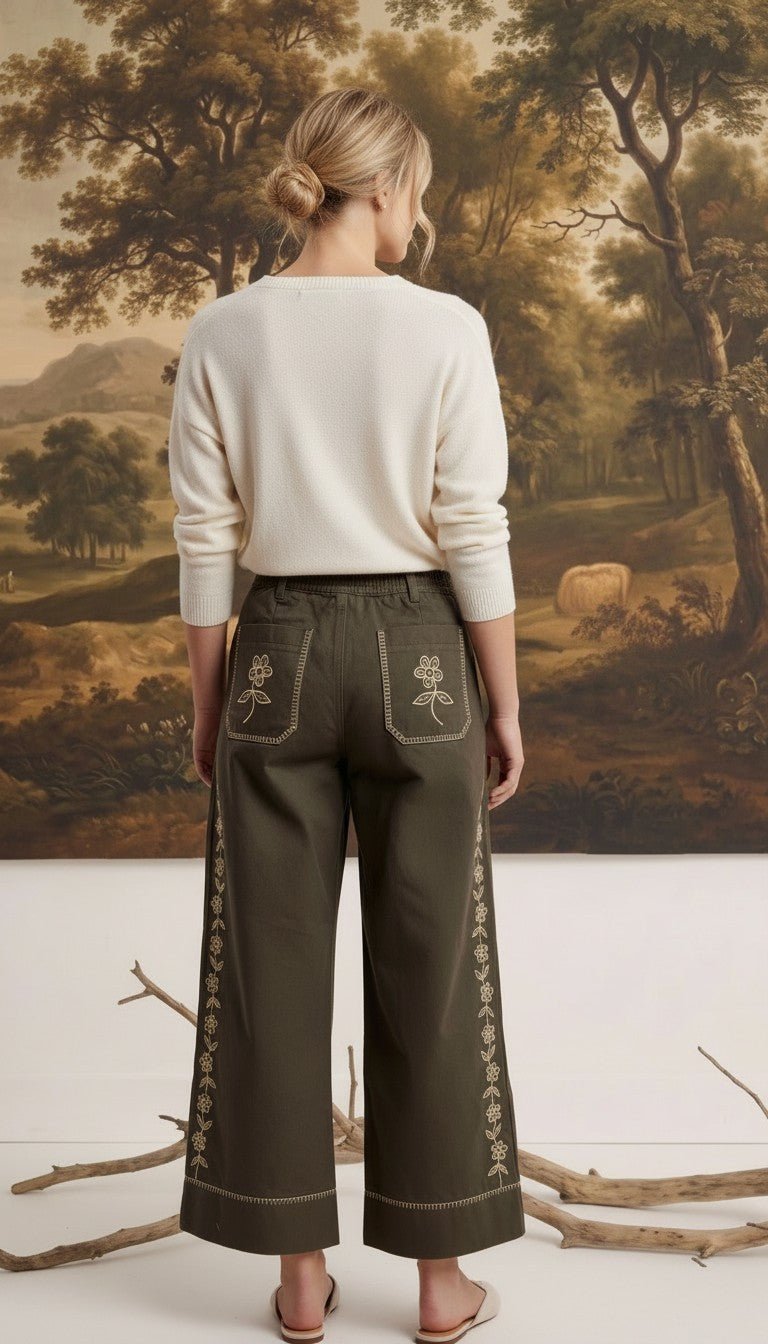 Model wearing graced olive green wide-leg cotton pants with floral embroidery, back view against landscape painting