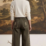 Model wearing graced olive green wide-leg cotton pants with floral embroidery, back view against landscape painting