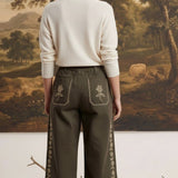 Model wearing graced olive green wide-leg cotton pants with floral embroidery, back view against landscape painting